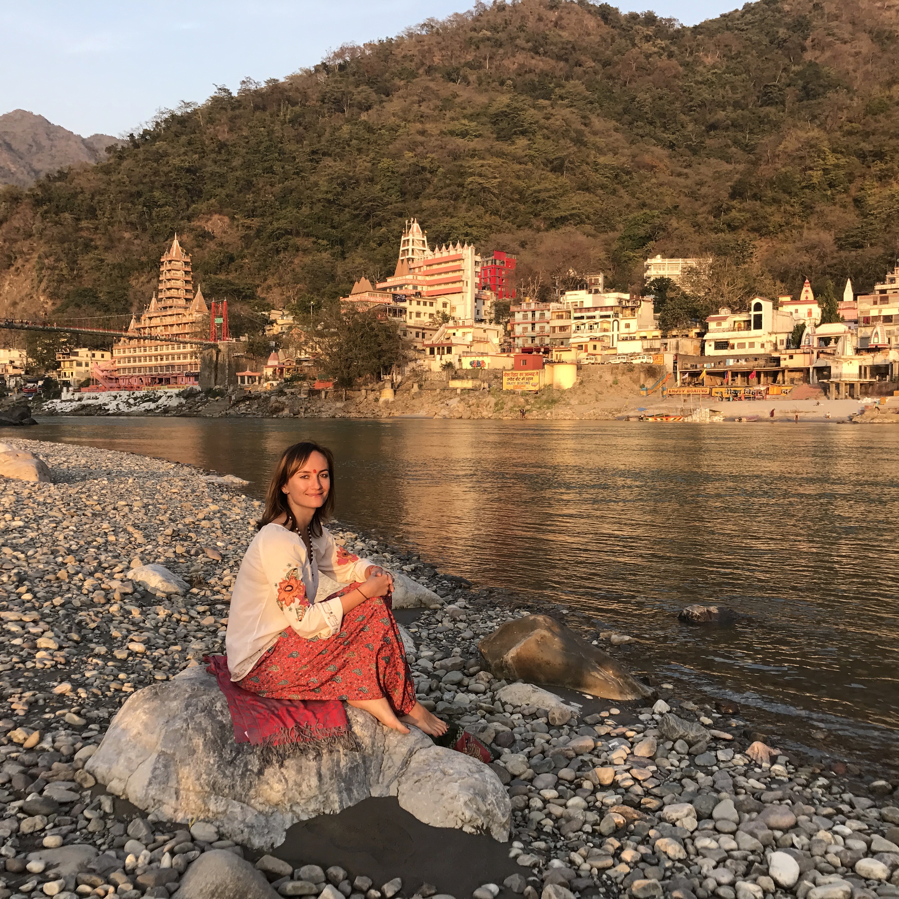 Zory sitting by the Ganges in Rishikesh, temples and mountains behind her at golden hour
